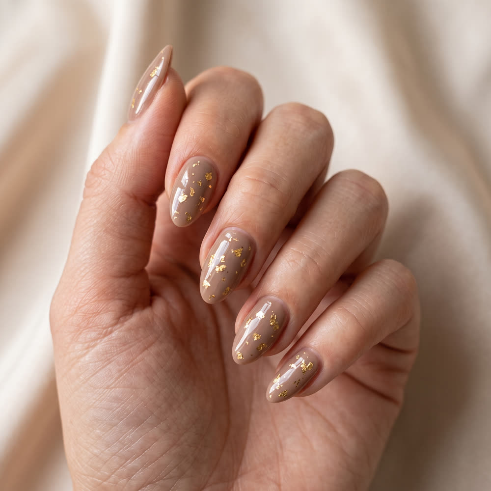 A hand on cream fabric with warm taupe nails carrying scattered gold flake
